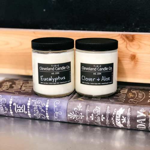 Two jars of Cleveland Candle Co. candles labeled "Eucalyptus" and "Clover & Aloe," placed on a decorative book.