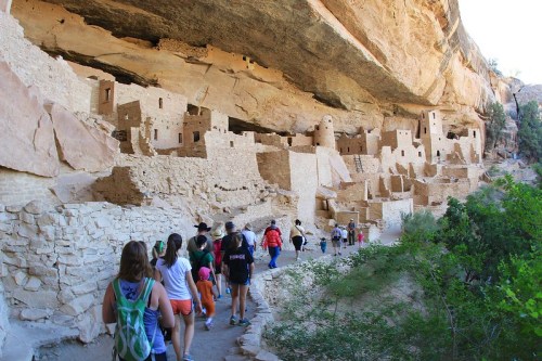 Visitors walk along a path near ancient cliff dwellings, surrounded by rocky terrain and greenery.