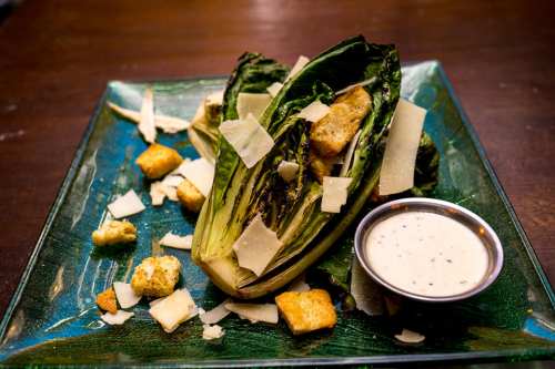 A plate of grilled romaine lettuce topped with croutons, shaved Parmesan, and a side of creamy dressing.