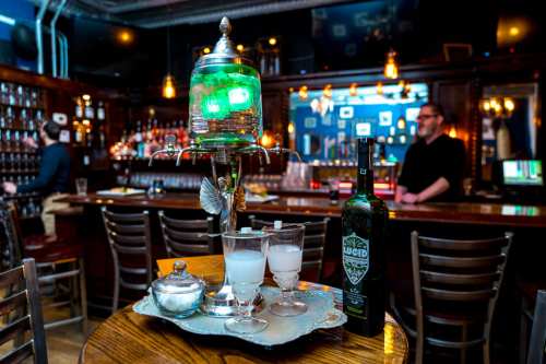 A stylish bar scene featuring a decorative absinthe fountain, glasses, and a bottle of Lucid absinthe on a table.