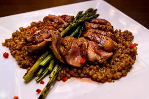 Sliced grilled meat atop a bed of lentils, garnished with asparagus and red pepper on a white plate.