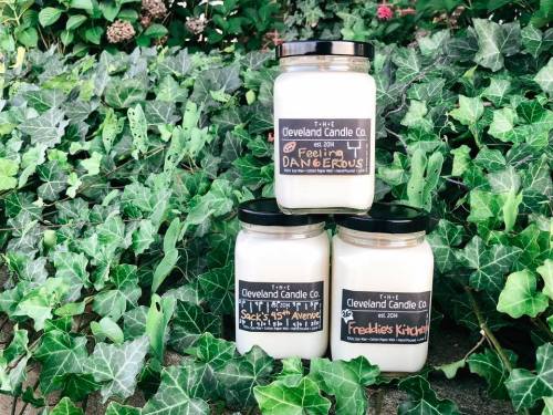 Three candle jars from The Cleveland Candle Co. sit against a backdrop of green ivy, labeled with unique scents.