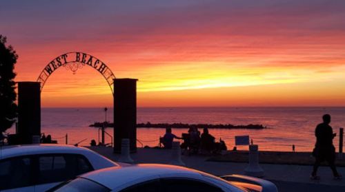 Sunset over the ocean with vibrant colors, featuring a "West Beach" arch and silhouettes of people enjoying the view.