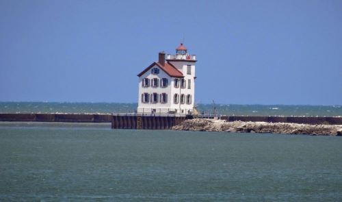 A white lighthouse with a red roof stands on a pier, surrounded by blue water and a clear sky.