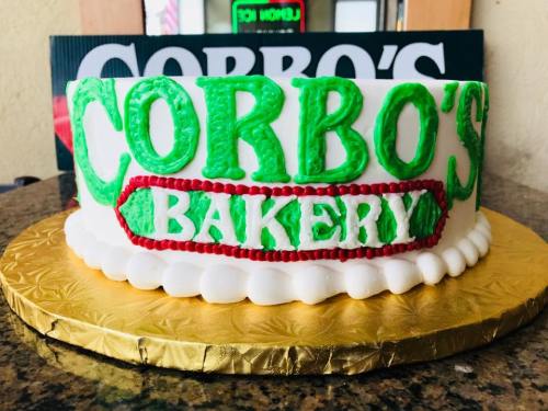 A round cake with green and red icing, featuring the text "Corbo's Bakery" in bold letters on a gold base.