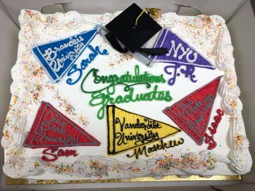 A graduation cake decorated with colorful pennants and names, celebrating multiple graduates from various universities.