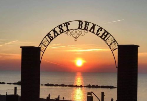 Sunset over a calm lake, framed by an archway that reads "East Beach," with silhouettes of people in the foreground.