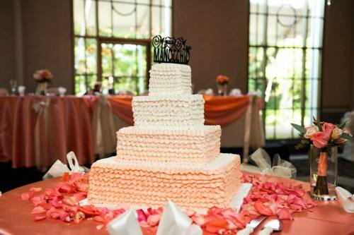 A three-tiered wedding cake with ruffled icing, topped with a black "Mr. & Mrs." cake topper, surrounded by rose petals.
