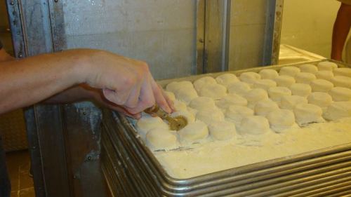 A hand using a dough cutter on a tray of round, uncooked dough pieces in a bakery setting.