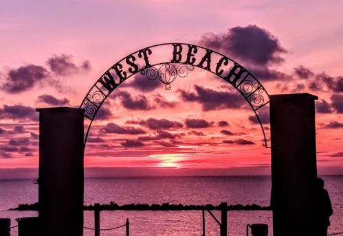 Silhouette of a "West Beach" arch against a vibrant sunset over the ocean, with clouds in the sky.