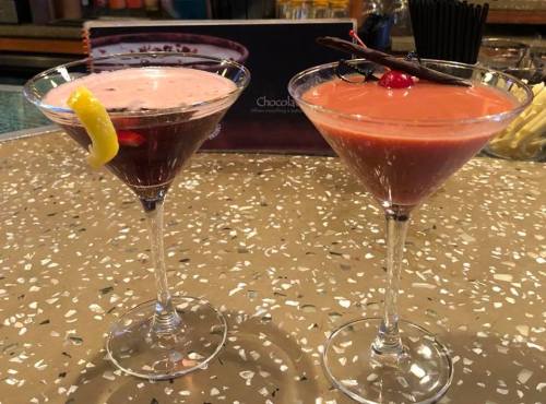Two colorful cocktails in martini glasses, one garnished with a lemon slice and cherry, the other with a cherry and cinnamon stick.