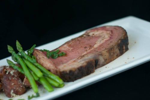 A slice of roast beef served with asparagus and roasted potatoes on a white plate against a black background.