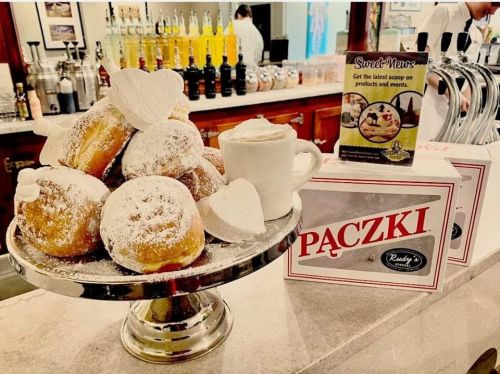 A silver platter piled with powdered sugar-dusted paczki, next to a cup and a sign about upcoming events.