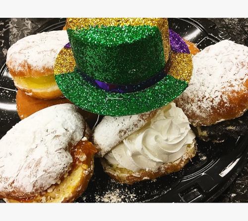A platter of powdered sugar donuts topped with a small green, purple, and gold glitter hat.