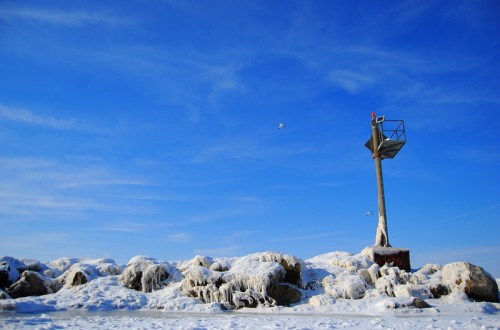 A frozen lighthouse on a rocky shore under a clear blue sky, with ice covering the rocks and a few birds flying nearby.