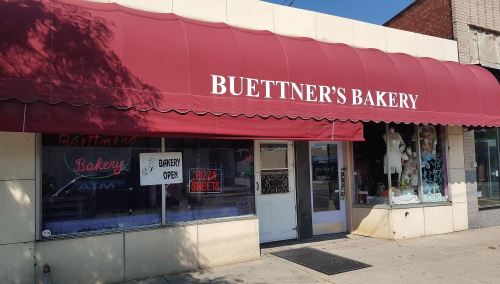 A storefront of Buettner's Bakery with a red awning and a sign indicating the bakery is open.