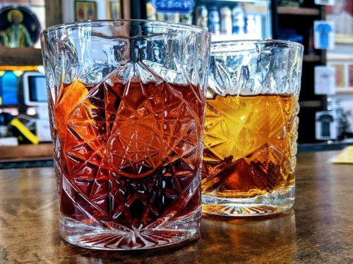 Two crystal glasses on a wooden bar, one filled with a dark red drink and the other with a golden amber beverage.