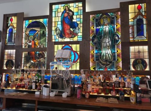 A bar with a variety of bottles and colorful stained glass windows depicting religious figures in the background.
