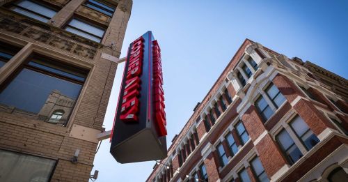 Sign for "Corner Alley" hanging above a street, with historic brick buildings and a clear blue sky in the background.