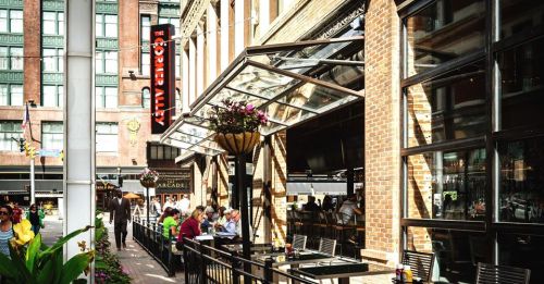 Outdoor dining area of a restaurant with large windows, flowers, and people enjoying their meals in a vibrant urban setting.
