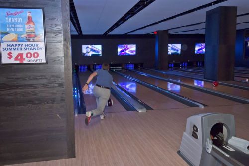 A person bowls in a modern bowling alley with screens and a sign advertising happy hour specials.