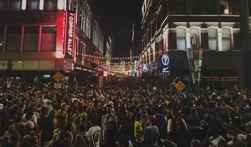 A large crowd gathers in a lively street at night, decorated with lights and signs, celebrating an event.