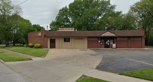 A brick building with a sign for a sausage shop, surrounded by trees and a parking lot.