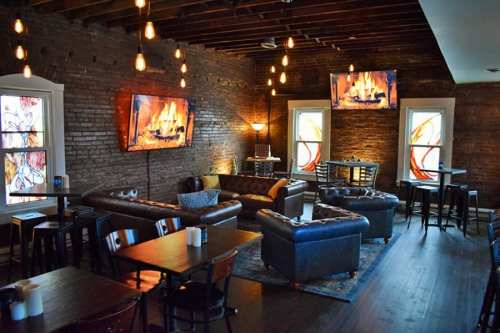 Cozy lounge with brick walls, leather sofas, and warm lighting, featuring TVs displaying a fireplace.