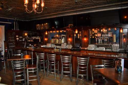 A cozy bar interior with wooden counters, metal chairs, and warm lighting, featuring shelves of drinks and a vintage ceiling.