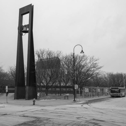 A large bell sculpture stands in a gray, urban setting with bare trees and a bus nearby.