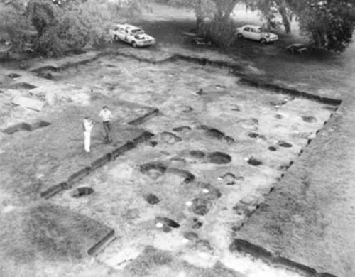 Aerial view of an archaeological dig site with two people examining the ground and parked cars in the background.
