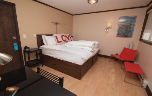 Cozy hotel room with a bed featuring large "LOVE" pillows, a desk, and a red chair against a beige wall.