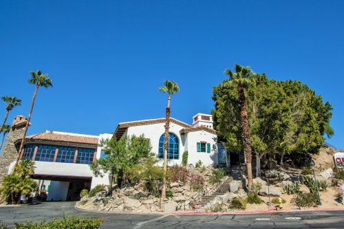 A large, white building with blue windows, surrounded by palm trees and rocky landscaping under a clear blue sky.