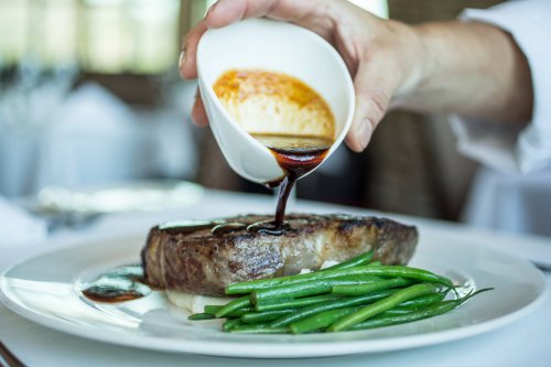 A hand pours sauce over a cooked steak, accompanied by green beans on a white plate.