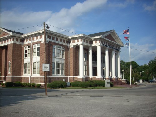 Historic brick building with columns, flags, and landscaped grounds, set against a blue sky with clouds.