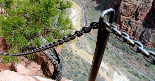 A metal chain along a rocky cliff edge, overlooking a winding river and lush greenery below.