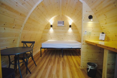 Cozy wooden interior of a small room featuring a bed, table, and air conditioning unit. Warm lighting enhances the space.