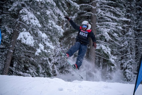 A snowboarder jumps off a snowy ramp, surrounded by trees, with snow spraying around them.