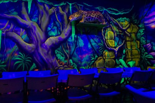 A vibrant mural of a jungle scene with a leopard on a ledge, illuminated by blue lighting, and empty chairs in the foreground.