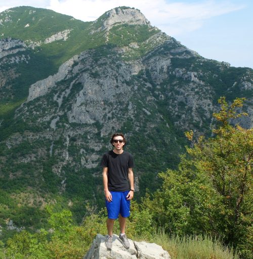 A person stands on a rock in front of a lush, green mountain landscape under a clear blue sky.