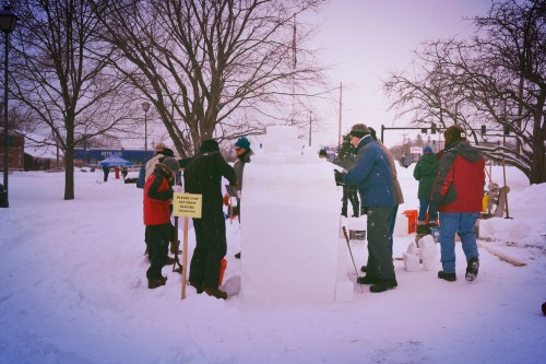 A group of people building a large snow sculpture in a snowy park, with trees and a sign nearby.