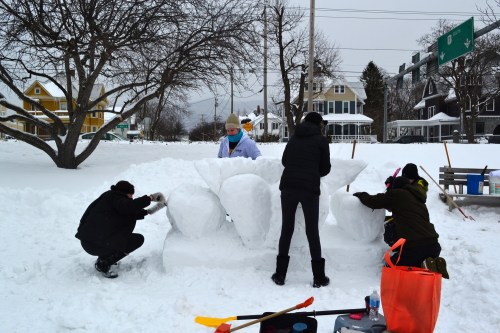 People sculpting a large snow structure in a snowy park, with trees and houses in the background.