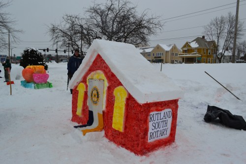 A colorful snow sculpture of a house with a sign reading "RUTLAND SOUTH ROTARY," surrounded by snow and winter scenery.