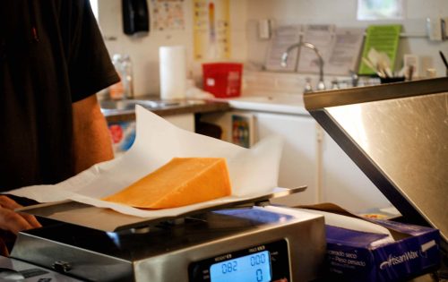 A block of cheese on a scale, with a person’s hand visible, in a kitchen setting.