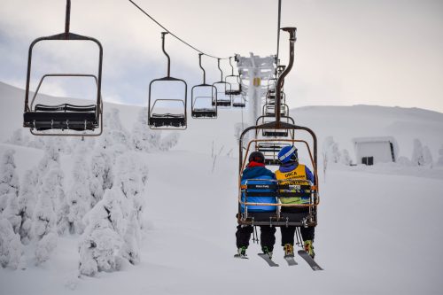 Two skiers on a chairlift surrounded by snow-covered trees and a cloudy sky.