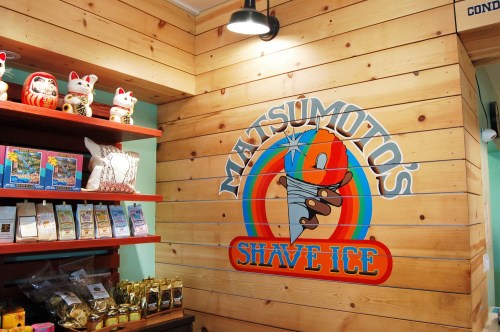Colorful wall mural of "Matsumoto's Shave Ice" with wooden paneling and various products displayed on shelves.