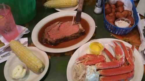 A table spread featuring prime rib, crab legs, corn on the cob, and rolls with butter and dipping sauce.