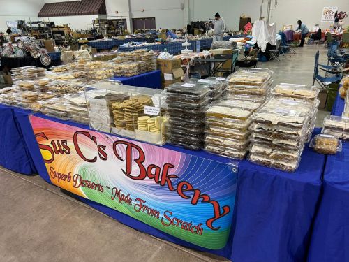 A bakery table filled with various desserts, featuring a colorful sign that reads "Sus C's Bakery - Super Desserts Made From Scratch."