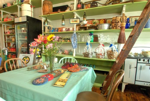 A cozy kitchen with a table set for dining, colorful decor, flowers, and shelves filled with vintage items and jars.