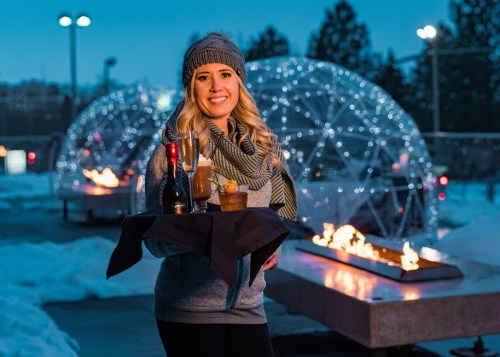 A smiling woman holds a tray of drinks in a snowy outdoor setting with illuminated igloos and fire pits in the background.
